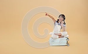 asian child sitting on suitcase pointing finger at copy space and look through binoculars isolated on color background.