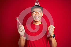 Asian chinese man on birthday celebration wearing funny hat over isolated red background screaming proud and celebrating victory