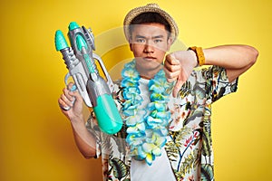 Asian chinese man wearing floral hawaian lei and water gun over isolated yellow background with angry face, negative sign showing