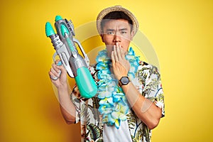 Asian chinese man wearing floral hawaian lei and water gun over isolated yellow background cover mouth with hand shocked with