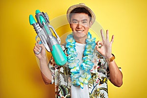 Asian chinese man wearing floral hawaian lei and water gun over isolated yellow background doing ok sign with fingers, excellent