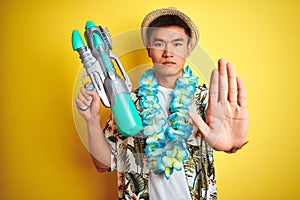 Asian chinese man wearing floral hawaian lei and water gun over isolated yellow background with open hand doing stop sign with