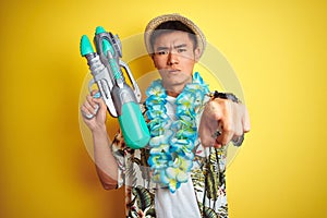Asian chinese man wearing floral hawaian lei and water gun over isolated yellow background pointing with finger to the camera and
