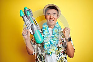 Asian chinese man wearing floral hawaian lei and water gun over isolated yellow background screaming proud and celebrating victory