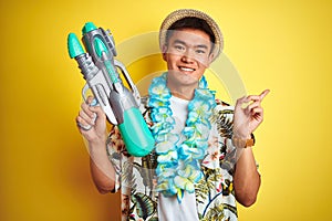 Asian chinese man wearing floral hawaian lei and water gun over isolated yellow background very happy pointing with hand and