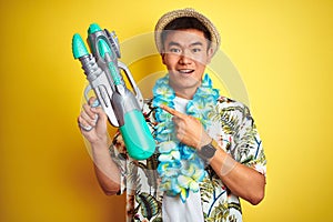 Asian chinese man wearing floral hawaian lei and water gun over isolated yellow background very happy pointing with hand and
