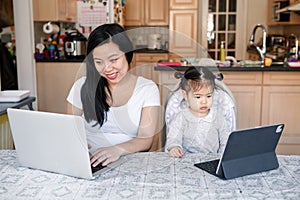 Asian Chinese mother with daughter baby working together on laptop, tablet. Workplace of freelancer woman with kid. Stay home mom