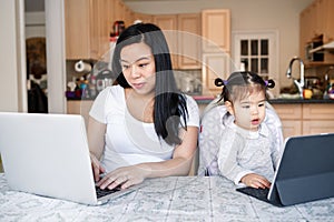 Asian Chinese mother with daughter baby working together on laptop, tablet. Workplace of freelancer woman with kid. Stay home mom