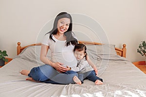 Asian Chinese pregnant woman with toddler girl sitting on bed at home. Girl daughter kid hugging mom belly. Mother and baby