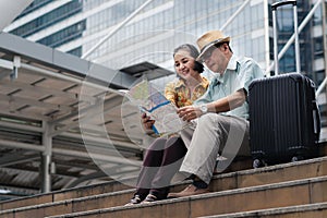 Asian chinese senior tourist couple with black luggage sitting together on stairway urban with map on hand after having city