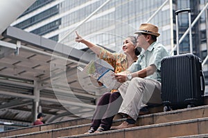 Asian chinese senior tourist couple with black luggage sitting together on stairway urban with map on hand after having city