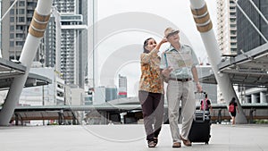 Asian chinese senior tourist couple with black luggage walking together in city with map on hand during having city sightseeing