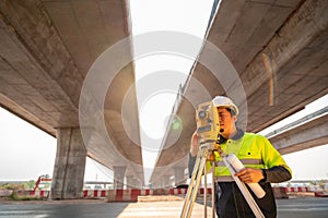 Asian civil engineer or surveyor holding a blueprint and making measuring under the expressway with theodolite on road works.
