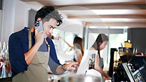 Asian coffee shop male employee accepts a pre-order on a mobile phone call while using a tablet computer in a cafe. Man