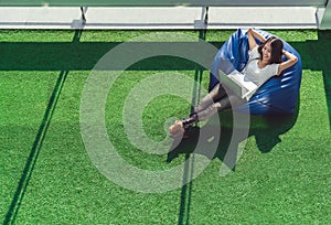 Asian college student or freelance woman using laptop computer, lying on bean bag in garden. University campus or park scene