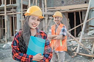 Asian construction worker woman smiles at the camera wearing a safety helmet against the background of a male contractor