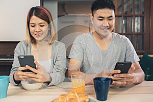 Asian couple distracted at table with newspaper and cell phone while eating breakfast.