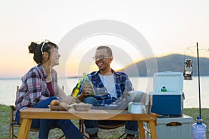 Asian couple drinking beer from bottle in their camping area with lake in the background during sunset