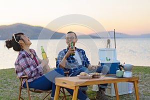 Asian couple drinking beer from bottle in their camping area with lake in the background during sunset