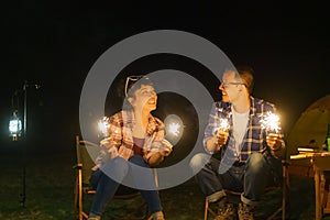 Asian couple is lighting sparkler fire at a campfire where they set up a tent to camp by the lake at night