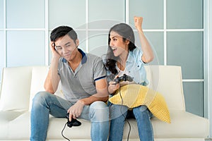 Asian couple man ans woman to playing video games with joysticks while sitting in sofa in living room at home and woman winning in