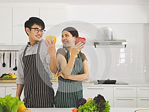 Asian couple preparing food in modern kitchen holding yellow and red capsicum smiling happily and looking each other