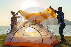 Asian couple preparing a tent to camping in the lawn with the lake in the background during sunset