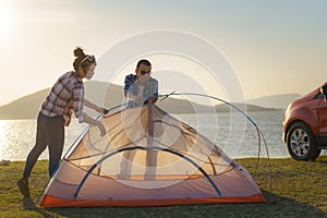 Asian couple preparing a tent to camping in the lawn with the lake in the background during sunset