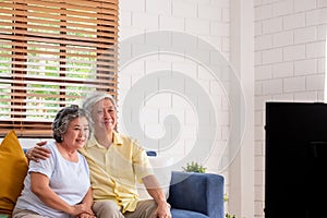 Asian couple senior sitting on sofa and  watching tv in living room at home.panning from blur foreground television