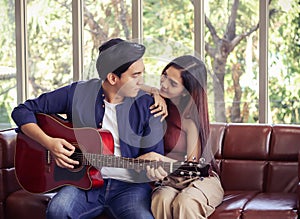 Asian couple sitting close together on couch in living room , a man playing guitar to his girlfriend and they are looking to each