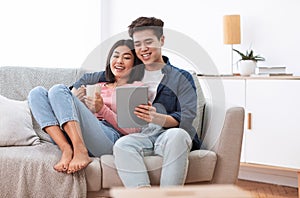 Asian Couple Using Digital Tablet Sitting On Sofa At Home