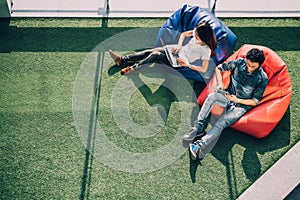 Asian couple using laptop notebook and digital tablet together in modern public park, sit on bean bag, top view with copy space