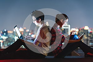 Asian couple using laptop and smartphone together, lean on each other on rooftop at night