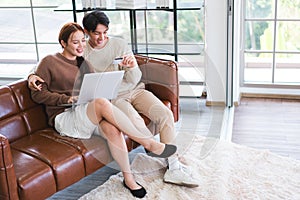 Asian couple wearing sweaters with a laptop, man uses a credit card to pay on the website in living room. E-commerce.