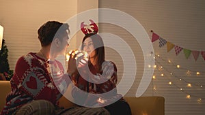 Asian couple wearing sweaters playing tangled led lights ball together on the sofa in the living room at home on Christmas eve