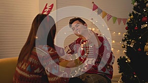Asian couple wearing sweaters playing tangled led lights together on the sofa in the living room at home on Christmas eve night.