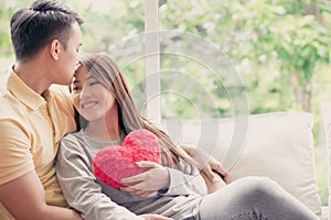 Asian Couples Sitting on the sofa In which women Holding a red heart And smiling happily. Concept of expressions of love and warmt