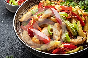 Asian cuisine, stir fry chicken, red paprika and zucchini with sesame and soy sauce in bowl. Black table background, top view,