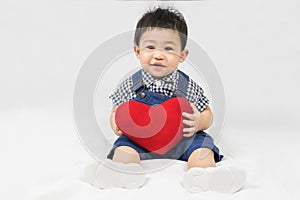 Asian cute baby boy about one year old in denim overalls and plaid shirts sitting and  smile with red heart shape pillow isolated