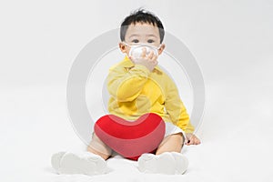 Asian cute baby boy about one year old in a protective mask and yellow shirt with red heart shape pillow, coronavirus and covid-19