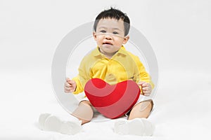 Asian cute baby boy about one year old in yellow shirt sitting and  smile with red heart shape pillow isolated on white Selective