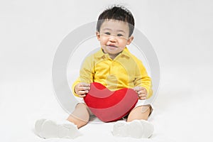 Asian cute baby boy about one year old in yellow shirt sitting and  smile with red heart shape pillow isolated on white Selective