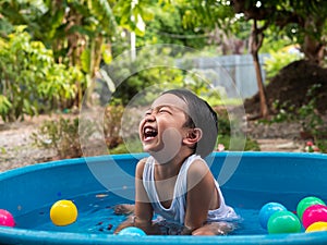 Asian cute child boy laughing while playing water in blue bowl with relaxing face and wet hair in rural nature.