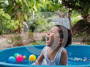 Asian cute child boy laughing while playing water in blue bowl with relaxing face and wet hair in rural nature with water splash.