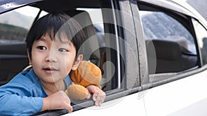 Asian cute child boy looking out car window with surprised face, hugging teddy bear with love in summer trip.