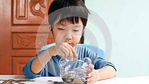 Asian cute child putting coin to transparent piggy bank by hand at home with happy face.