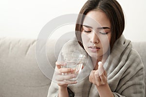 Asian Cute of  girl having headache and high temperature from illness holding pill of medicine and water sitting on sofa at home,