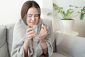 Asian Cute of  girl having headache and high temperature from illness holding pill of medicine and water sitting on sofa at home,