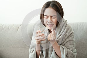 Asian Cute of  girl having headache and high temperature from illness holding pill of medicine and water sitting on sofa at home,