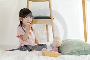 Asian cute little girl eating snack while sitting in a blanket fort in living room at home for perfect hideout away from their